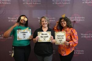 Three women stand smiling in front of a Western University of Health Sciences backdrop, each holding signs with zest-themed messages and wearing lemon-themed props.