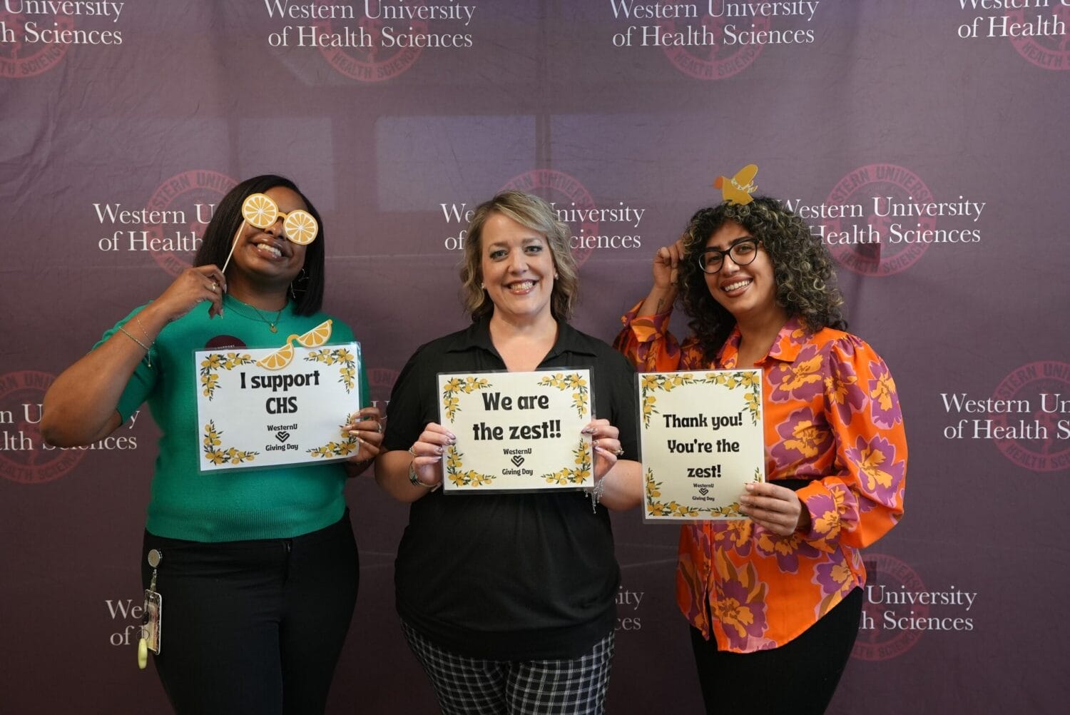 Three women stand smiling in front of a Western University of Health Sciences backdrop, each holding signs with zest-themed messages and wearing lemon-themed props.