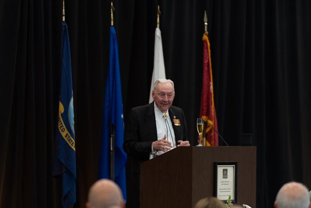 An elderly man in a suit speaks at a podium with military flags behind him at an indoor event.