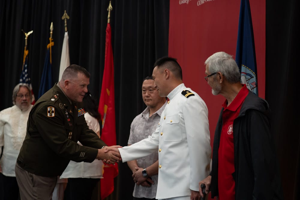 A uniformed officer shakes hands with a man in a white naval uniform on stage, while three other men stand nearby in front of flags and a red backdrop.