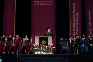 A speaker in academic regalia stands at a podium on stage during a graduation ceremony at Western University of Health Sciences, surrounded by faculty and staff in formal attire.