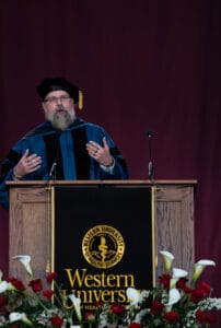 A speaker in academic regalia stands at a podium with the Western University of Health Sciences logo, addressing an audience; flowers decorate the front of the stage.