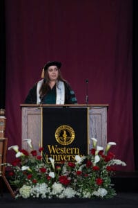 A woman in graduation attire stands at a podium with a "Western University" banner, delivering a speech. The stage is decorated with red and white flowers.