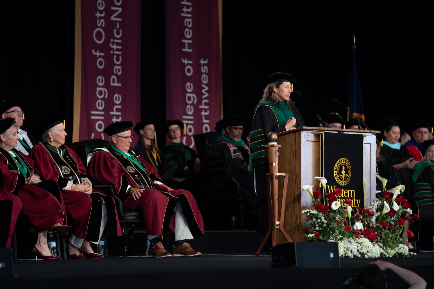 A woman in academic regalia speaks at a podium during a graduation ceremony, with faculty and officials seated on stage behind her.