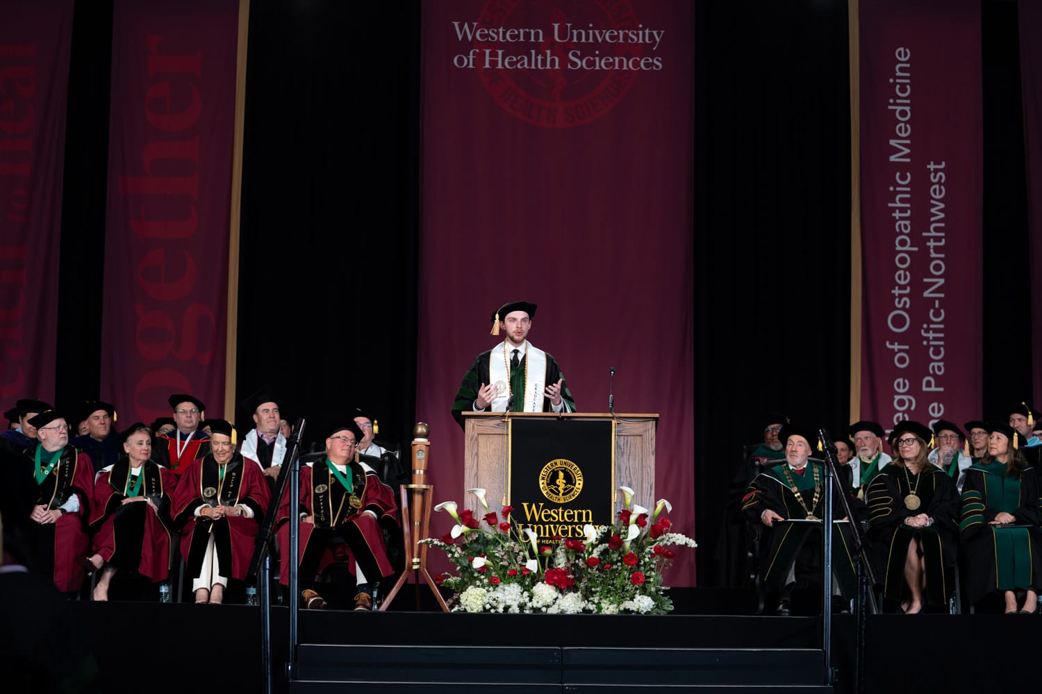 A speaker in academic regalia addresses an audience from a podium at a Western University of Health Sciences graduation ceremony, with faculty and officials seated on stage.