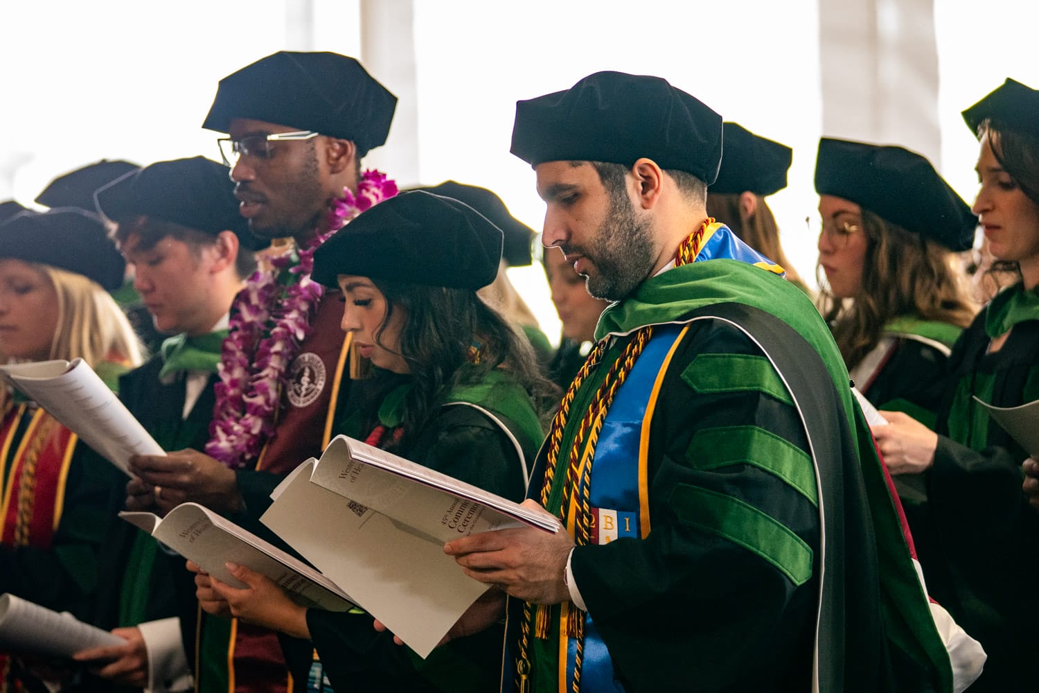 A group of graduates in caps and gowns stands in a row, reading from programs during a commencement ceremony.