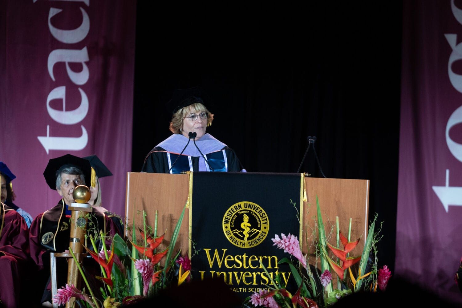 A person in academic regalia speaks at a podium with the Western University logo during a graduation ceremony, with banners and flowers in the background.