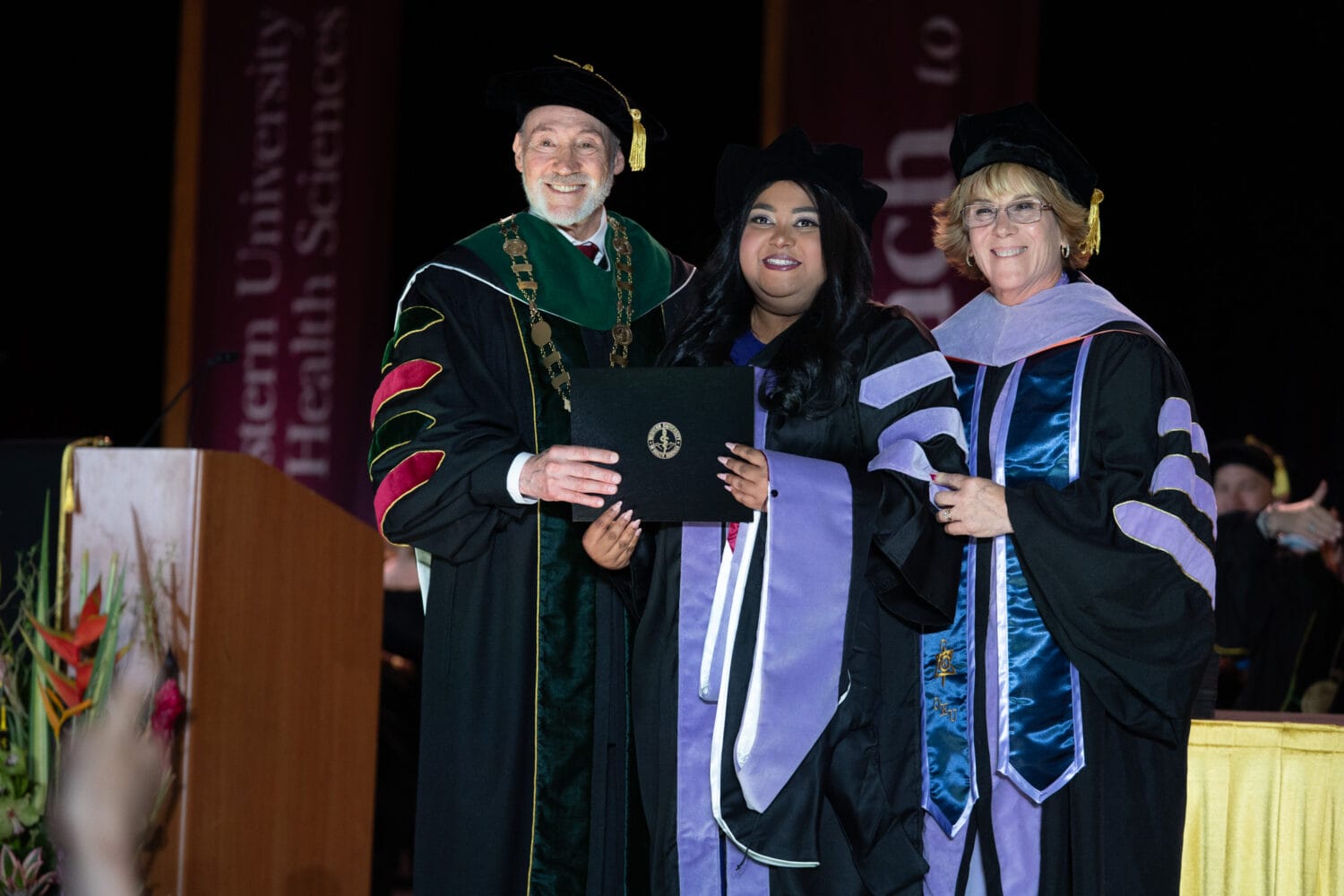 Three people in academic regalia pose on stage at a graduation ceremony. The person in the middle holds a diploma.