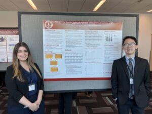 Two people stand in front of a research poster on orthodontic nanoparticles at an academic conference. Both are dressed in business attire and smiling at the camera.