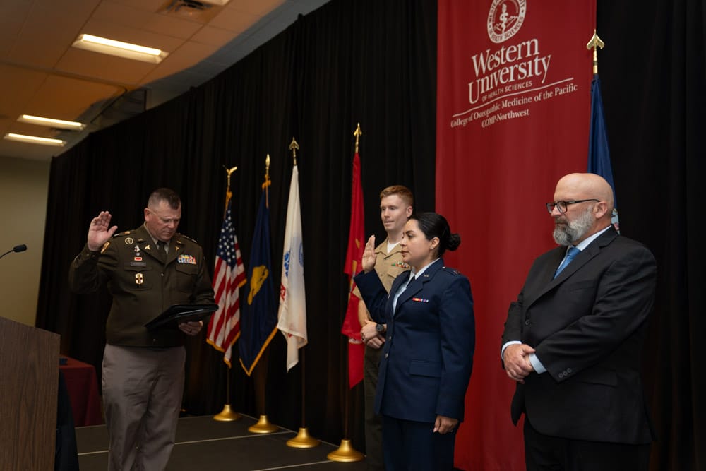 A military officer administers an oath to two uniformed individuals as another person in a suit stands nearby. Flags and a Western University banner are in the background.