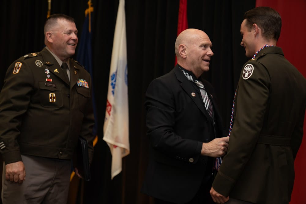 Three men stand together at a formal event; one man awards a cord to another in military dress while a second uniformed man looks on, smiling.