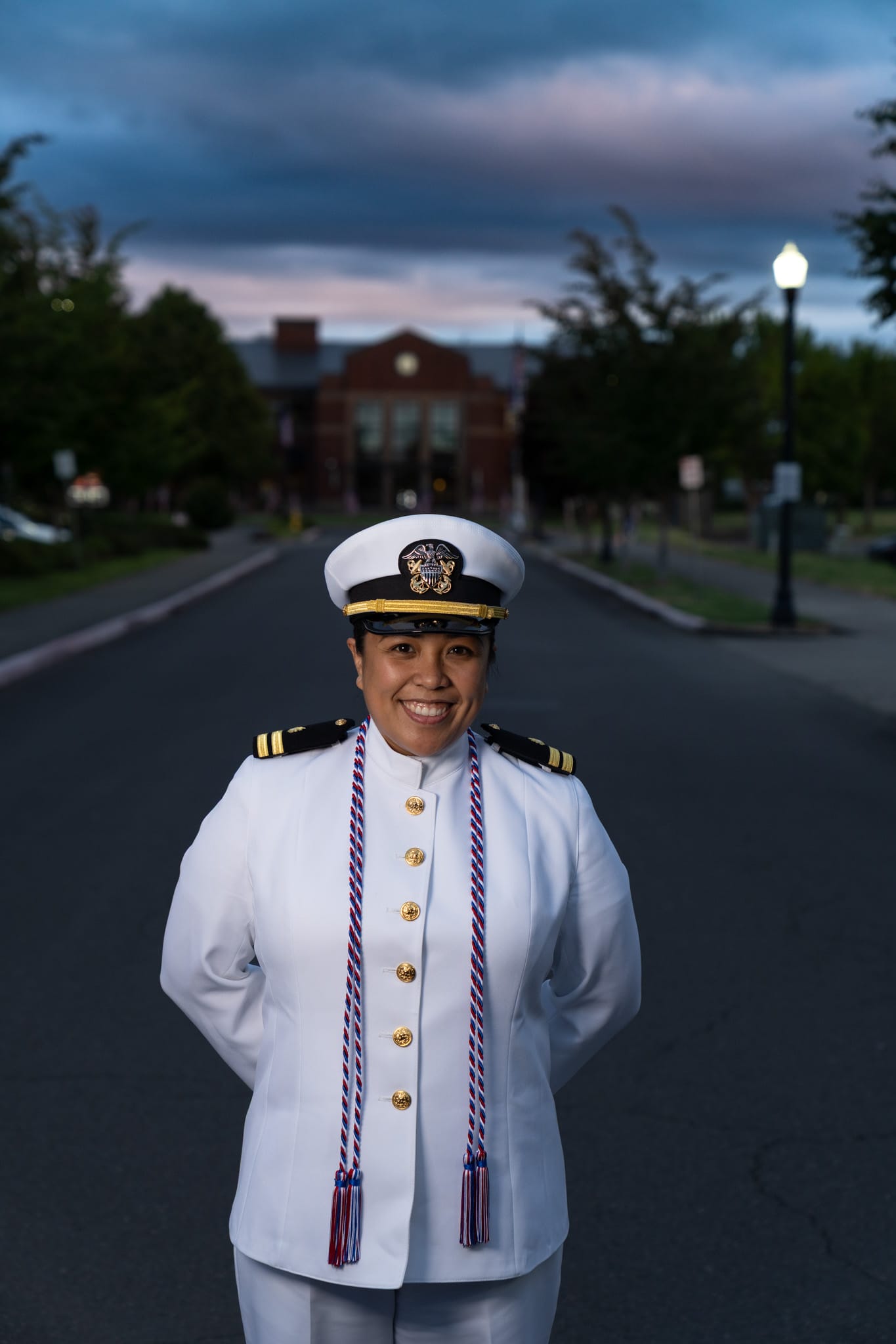 A person in a white military dress uniform with a cap stands smiling on a street with trees and a building in the background at dusk.