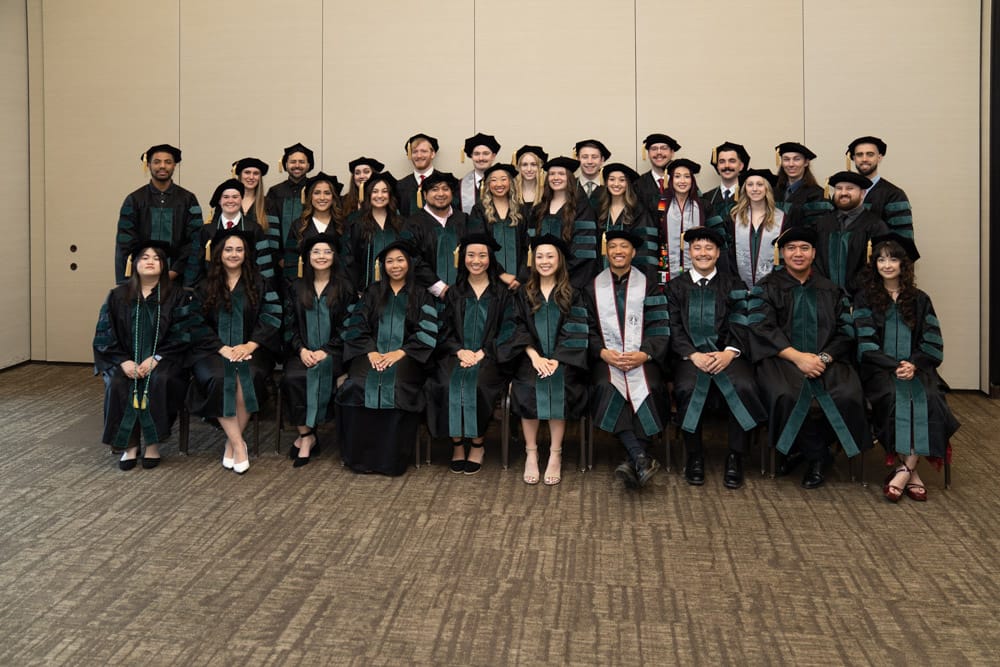 A group of graduates in caps and gowns pose for a photo indoors against a beige wall, arranged in three rows.