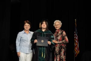 A graduate in cap and gown holds a diploma while standing between two older women, with a U.S. flag in the background.