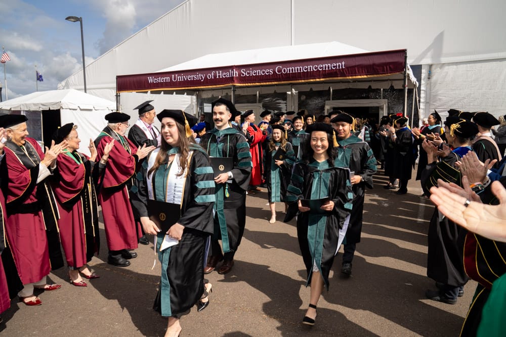 Graduates in academic regalia walk past applauding faculty outside a tent with a banner reading "Western University of Health Sciences Commencement.