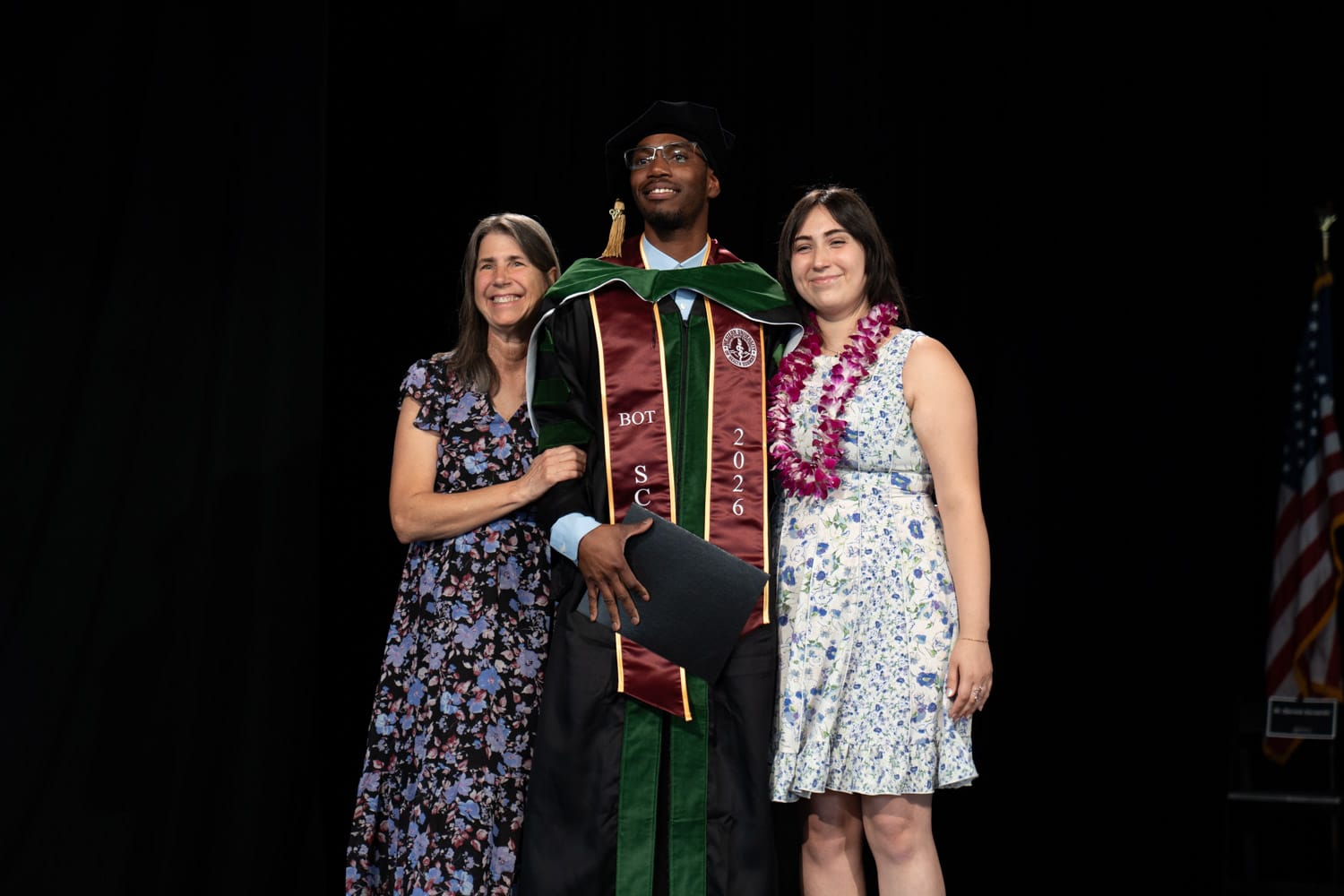 A graduate in academic regalia stands holding a diploma, flanked by two women in floral dresses, against a black background with a partial US flag visible.