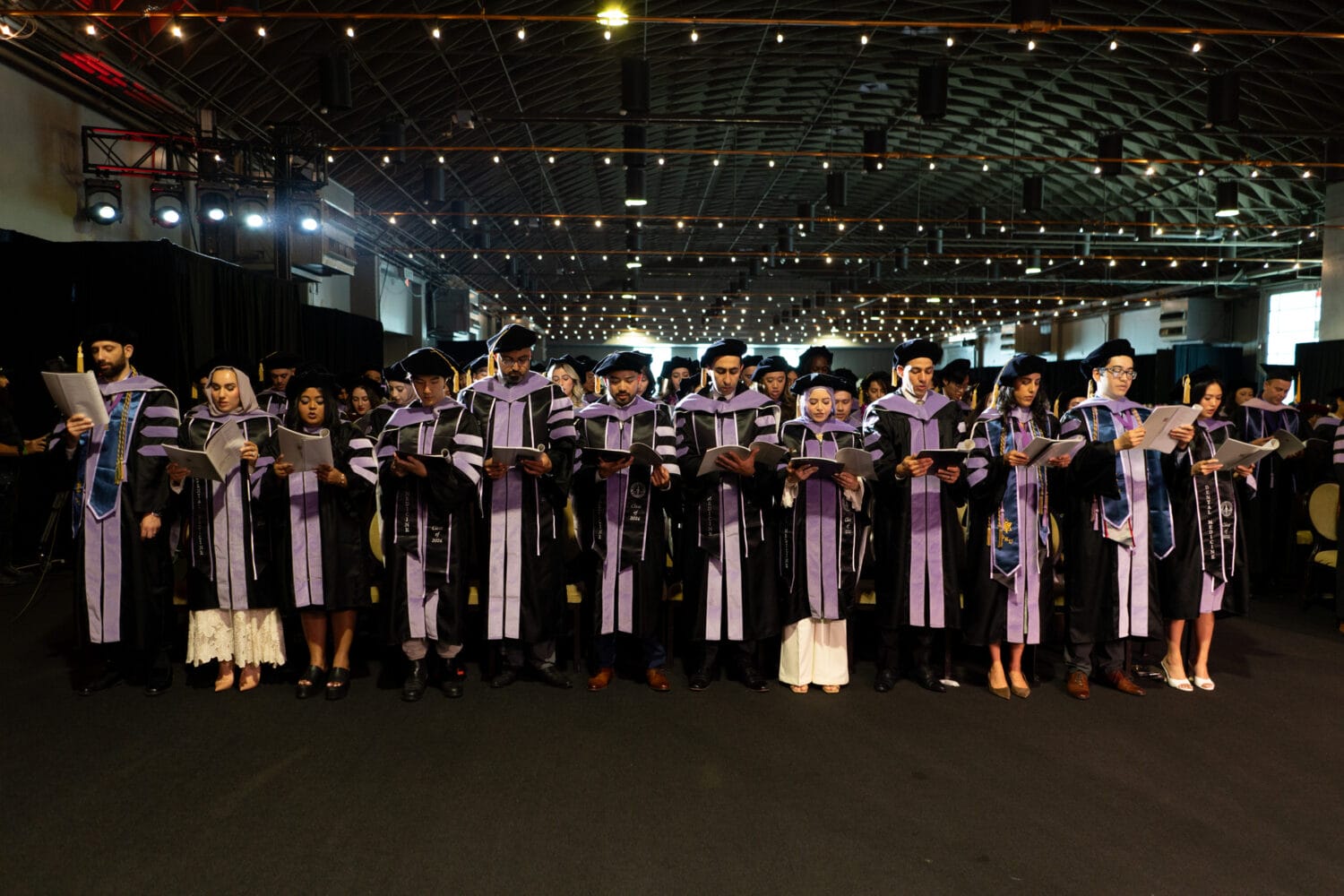 A group of graduates in caps and gowns stand in a row, holding papers and reading during a commencement ceremony in a large indoor venue.