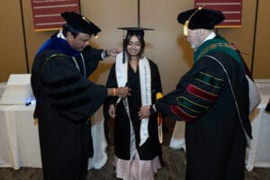 Two faculty members in academic regalia place an honors sash on a student in a graduation gown during a ceremony.