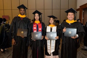 Four graduates in caps and gowns stand indoors, holding their diplomas and smiling at the camera.
