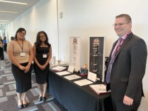 Three people stand next to a display table with diagrams, models, and documents in a conference setting.