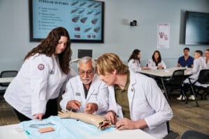 Three medical professionals in white coats practice suturing on a mannequin arm in a classroom, with a sutures diagram displayed on a screen in the background.