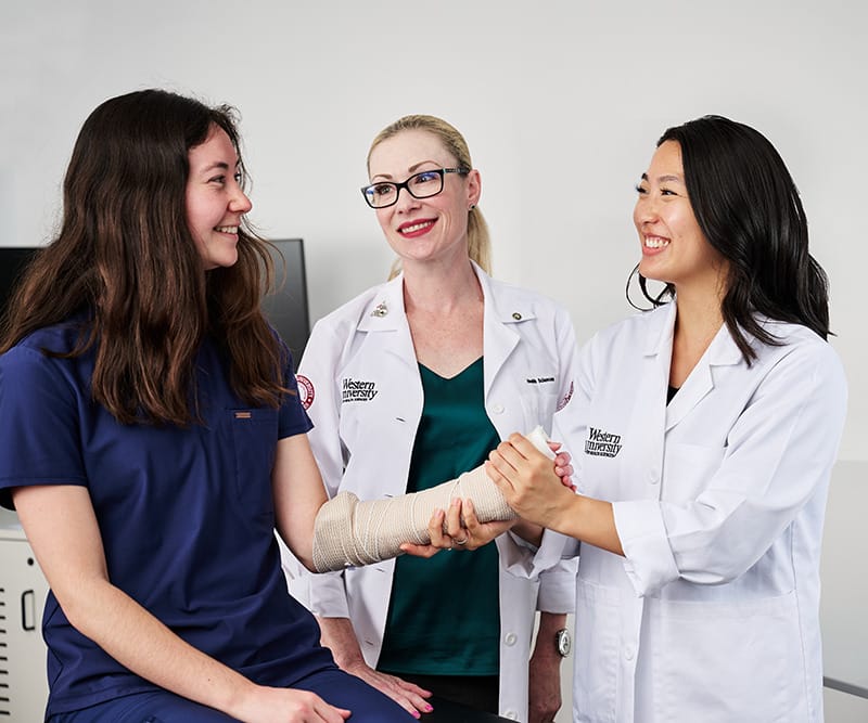 Two healthcare professionals examine the bandaged arm of a smiling patient who is wearing navy scrubs.