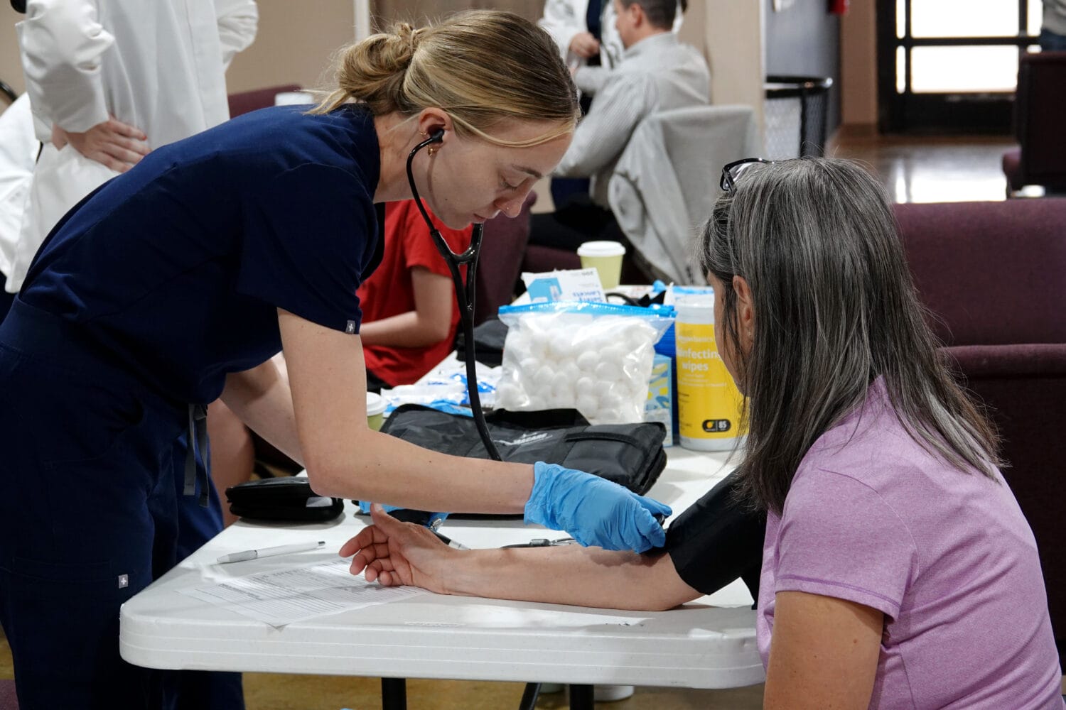 A healthcare worker in blue scrubs measures a seated woman's blood pressure at a table with medical supplies on it.