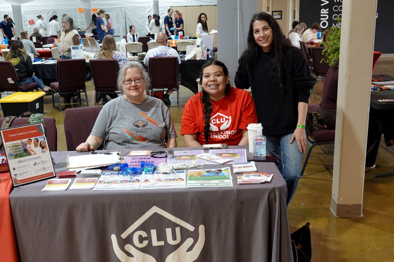 Three women sit and stand behind a table with informational materials at an indoor event, representing an organization called CLU. Other tables and attendees are visible in the background.