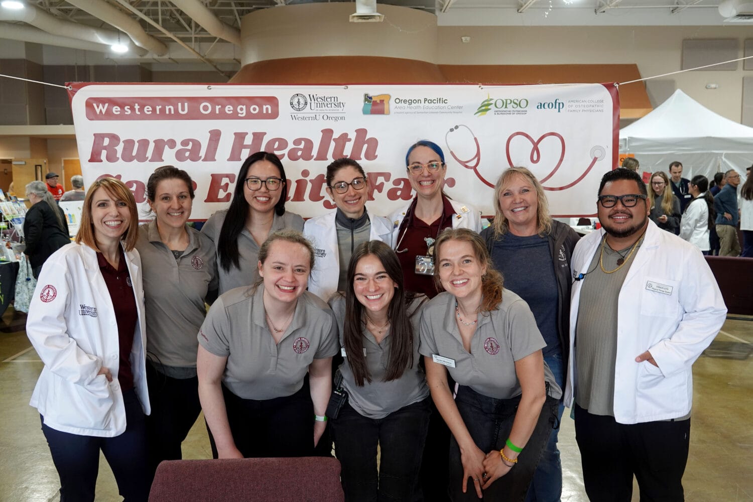 A group of people in matching polos and lab coats pose and smile in front of a "Rural Health Care Exploration Fair" banner at an indoor event.
