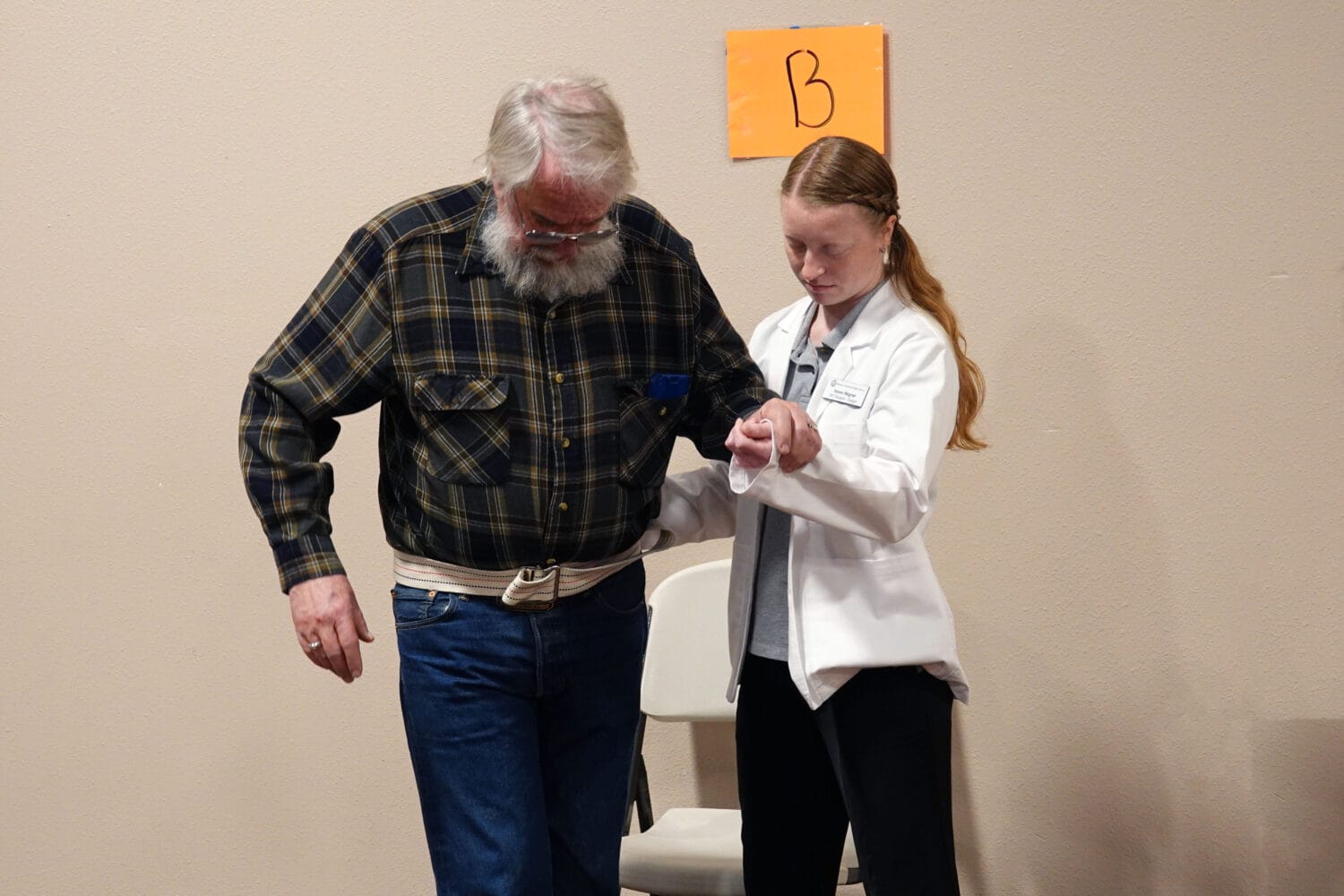 A woman in a white coat assists an older man with a gait belt as he stands next to a chair, with an orange sign marked "B" on the wall behind them.