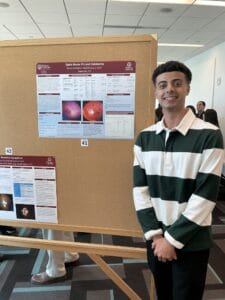 A young man stands and smiles in front of a science poster about optic nerve pathology displayed on a corkboard at an indoor event.