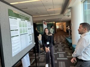A woman presents a research poster to a man in a hallway with several other posters and people visible in the background.