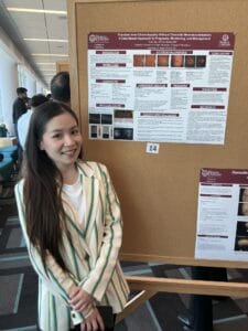 A woman in a striped blazer stands and smiles in front of a scientific poster presentation on a corkboard at a conference.