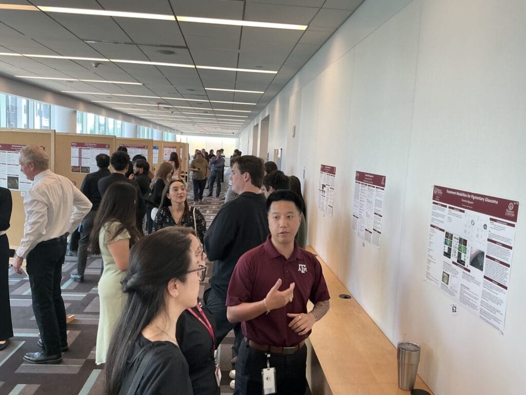 People view and discuss research posters displayed on the wall at an indoor academic event; one man in a maroon shirt speaks to an attendee in the foreground.