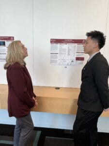 Two people in business attire stand and talk in front of a scientific research poster displayed on a white wall at an academic event.
