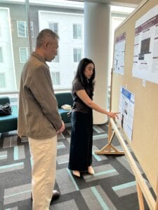 A woman points to a poster on a bulletin board while speaking to a man; they are indoors with windows and seating in the background.