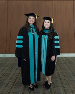 Two women wearing academic regalia, including doctoral gowns and caps, stand side by side and smile in front of a wooden wall.