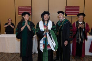 A graduate in academic regalia smiles and claps, surrounded by faculty in gowns at a graduation ceremony.