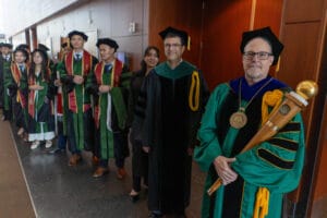 A group of graduates in green academic gowns stand in a hallway with two faculty members in black gowns, one holding a ceremonial mace.