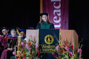 A speaker in academic regalia stands at a podium with a Western University of Health Sciences sign during a graduation ceremony, with faculty seated in the background.