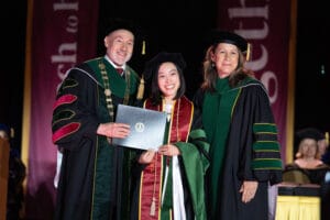 Three people in academic regalia pose on stage; the graduate in the center holds a diploma while smiling, flanked by two officials during a graduation ceremony.