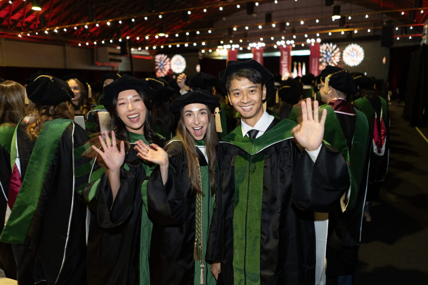 Three graduates in academic regalia smile and wave at the camera during a graduation ceremony in a large indoor venue.