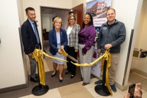 Five people stand together at a ribbon-cutting ceremony indoors, with one person using large scissors to cut a yellow ribbon in front of a doorway.