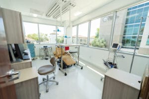 A modern dental clinic room with a dental chair, equipment, cabinets, and large windows overlooking a university building.