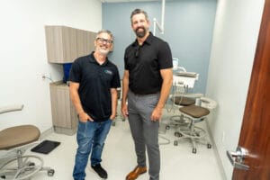 Two men stand and smile in a modern dental office, surrounded by dental chairs and equipment.