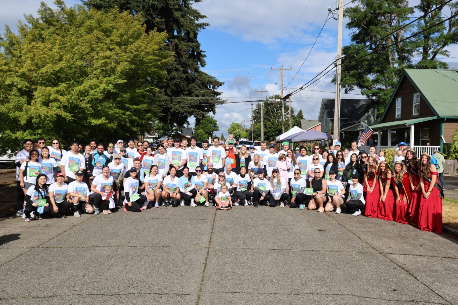 A large group of people pose for a group photo outdoors on a sunny day; most are wearing matching white T-shirts, with a few people in red costumes standing at the right.
