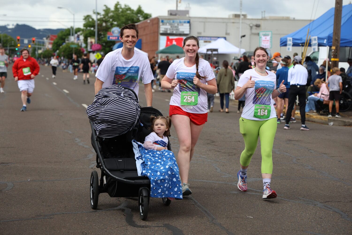 Three adults run in a race on a city street, two wearing race bibs, one pushing a stroller with a young child covered by a blanket. Other runners and tents are visible in the background.
