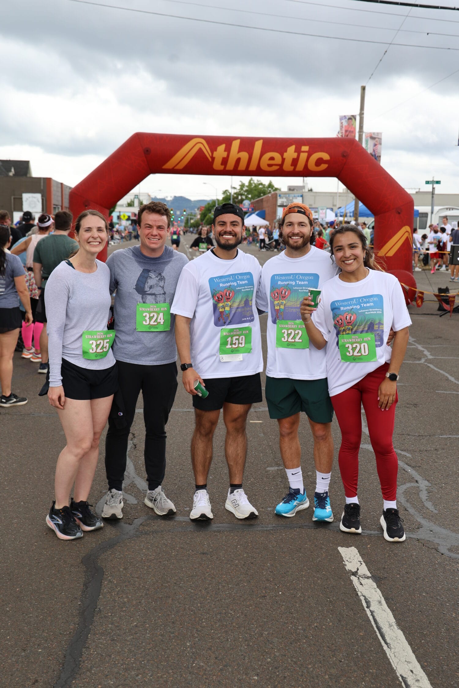 Five runners pose together in front of a red race arch, wearing race bibs and athletic clothing, at an outdoor running event.