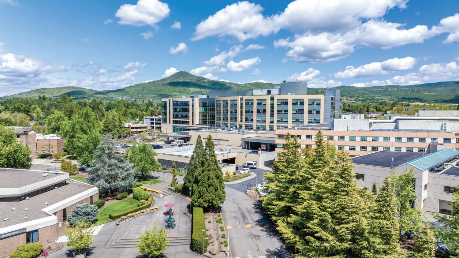 A modern hospital complex surrounded by trees and greenery, with a mountain and blue sky in the background.