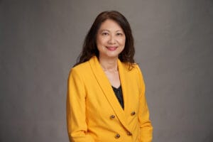 A woman with long brown hair wearing a bright yellow blazer over a black top poses in front of a plain gray background.